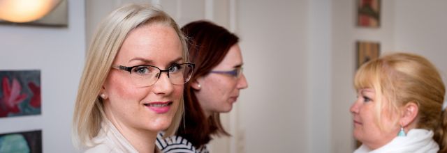 Eine Frau mit blondem Haar blickt in die Kamera, zwei weitere Frauen unterhalten sich im Hintergrund.